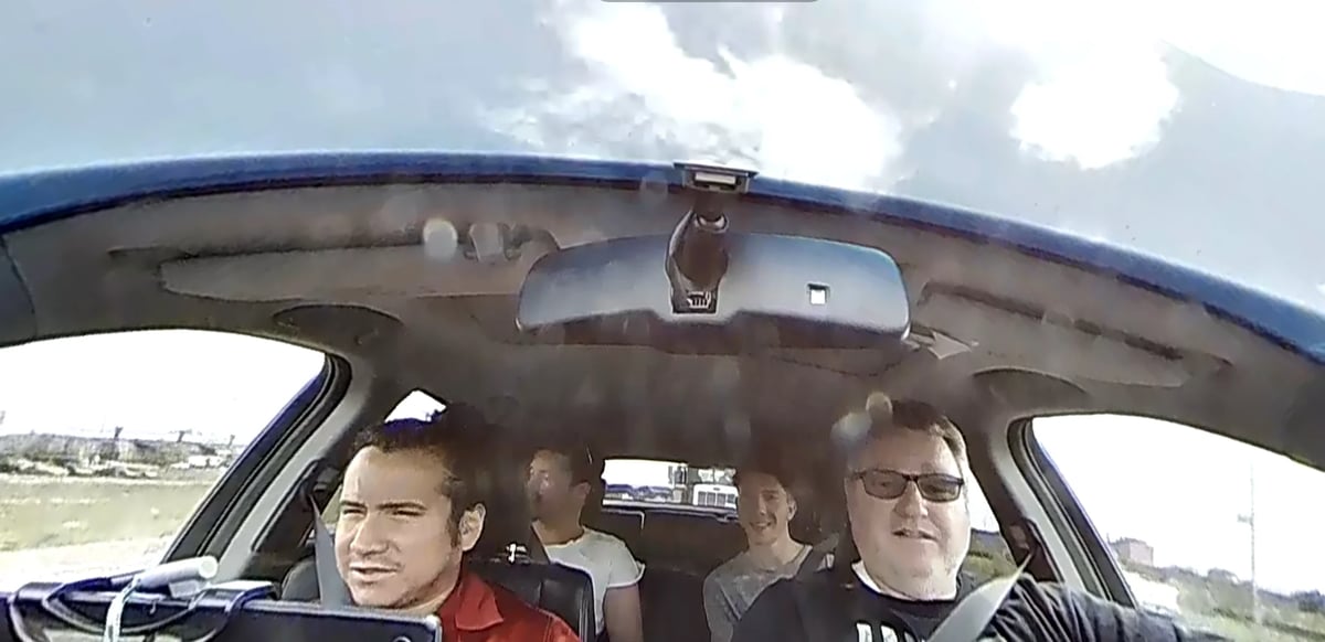 Three men sitting in the front seat of a car with blue trim, viewed from inside the vehicle.