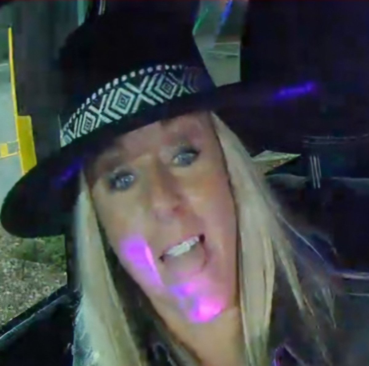 Woman with blonde hair wearing a black cowboy hat with geometric pattern band, smiling at camera with purple stage lighting effects
